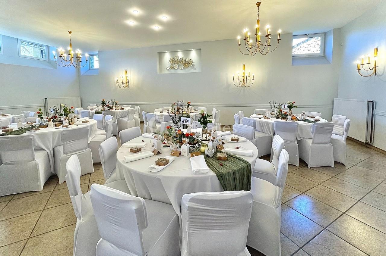 Salle de réception lumineuse avec tables rondes décorées de nappes blanches, centres de table floraux et chandeliers.