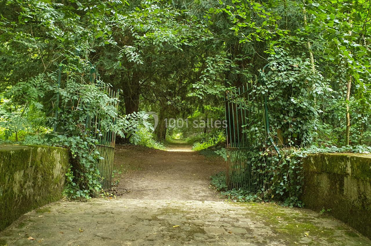 Chemin forestier traversant une arche naturelle de végétation, encadré par une barrière métallique et un pont en pierre.