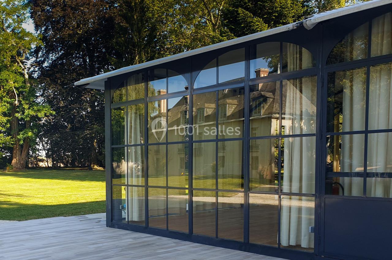Verrière moderne donnant sur un jardin arboré, reflétant une maison ancienne sous un ciel dégagé.