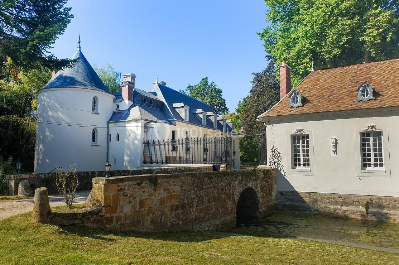Petit château avec tourelle et dépendance, entouré d'arbres, avec un pont en pierre traversant un cours d'eau.