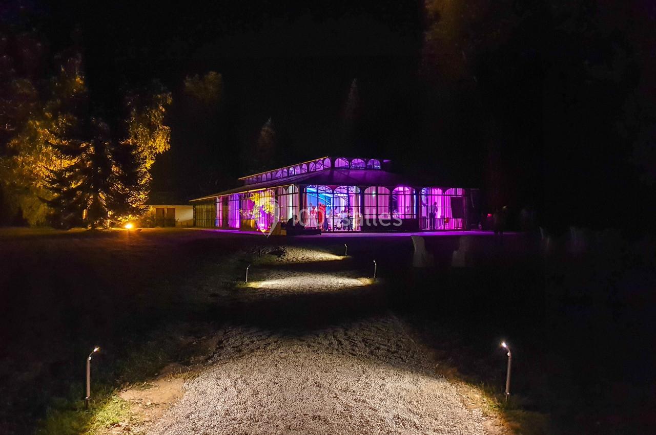 Chemin éclairé menant à un bâtiment avec façade vitrée illuminée de lumières colorées dans un cadre nocturne.