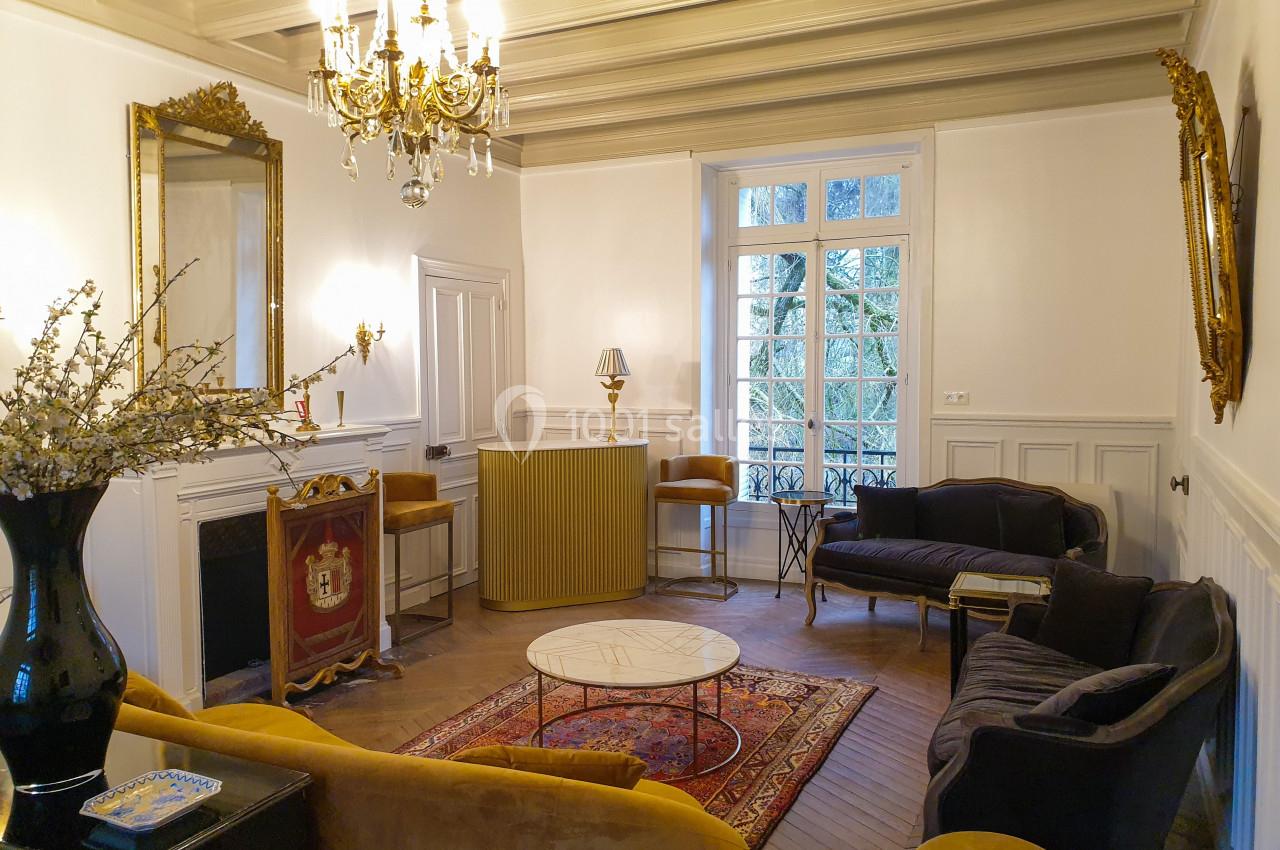 Salon élégant avec mobilier vintage, cheminée, lustre doré, miroirs et grandes fenêtres laissant entrer la lumière naturelle.