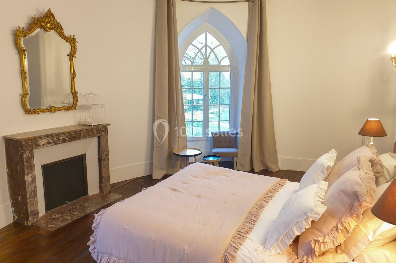 Chambre lumineuse avec lit double, cheminée en marbre, miroir doré et grande fenêtre en arc avec rideaux beiges.