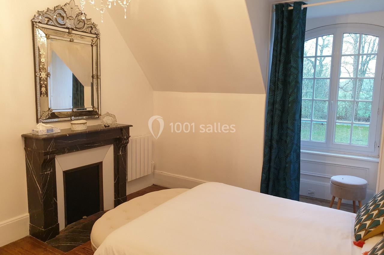 Chambre lumineuse avec un lit, une cheminée décorative, un miroir, des rideaux bleus et une fenêtre donnant sur un jardin.