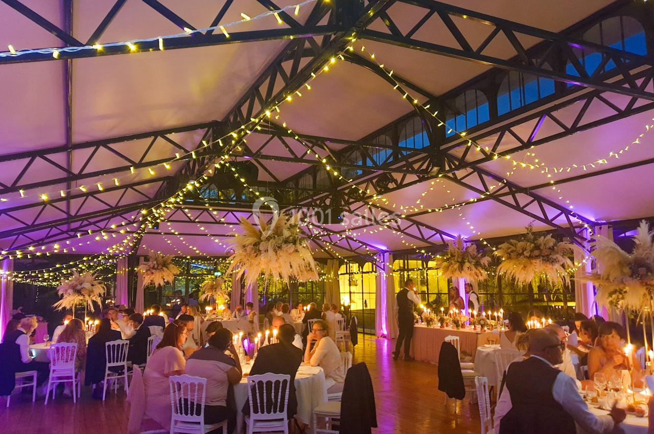 Salle de réception décorée avec des guirlandes lumineuses, des tables dressées et des invités assis lors d'un événement.
