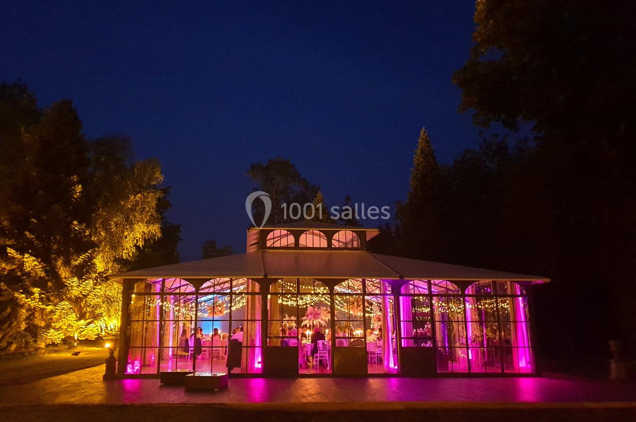 Salle de réception vitrée illuminée en rose, entourée d'arbres et visible de nuit sous un ciel sombre.