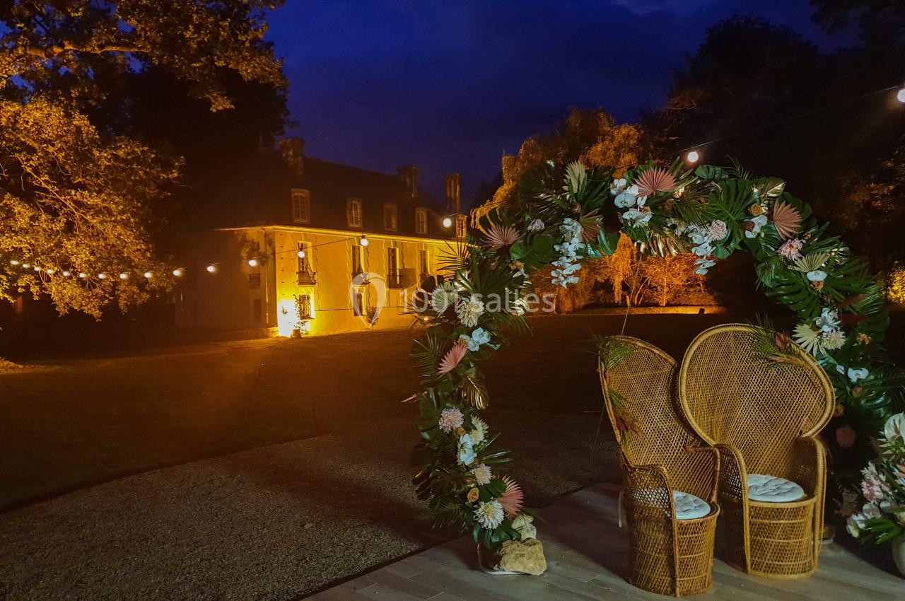 Arc floral illuminé avec deux fauteuils en rotin devant un bâtiment historique éclairé, de nuit.