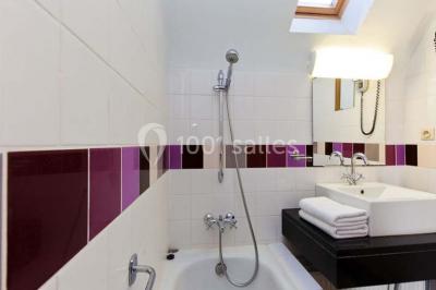 Salle de bain avec baignoire, carrelage blanc et violet, lavabo avec miroir, serviettes pliées et lumière naturelle.