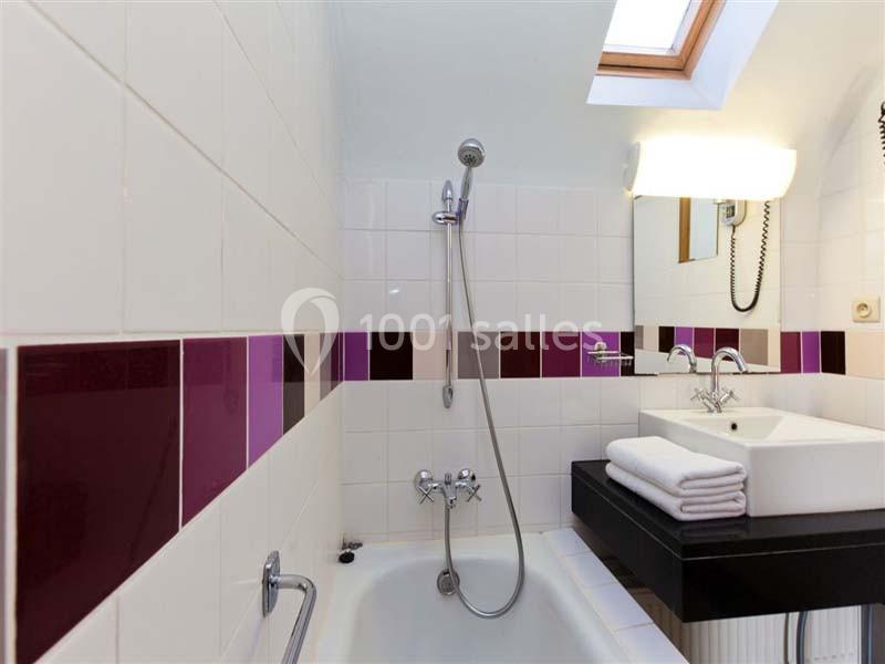 Salle de bain avec baignoire, carrelage blanc et violet, lavabo avec miroir, serviettes pliées et lumière naturelle.