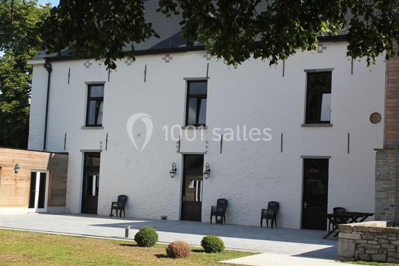 Façade d'un bâtiment blanc avec fenêtres noires, terrasse pavée, chaises et arbustes au premier plan.