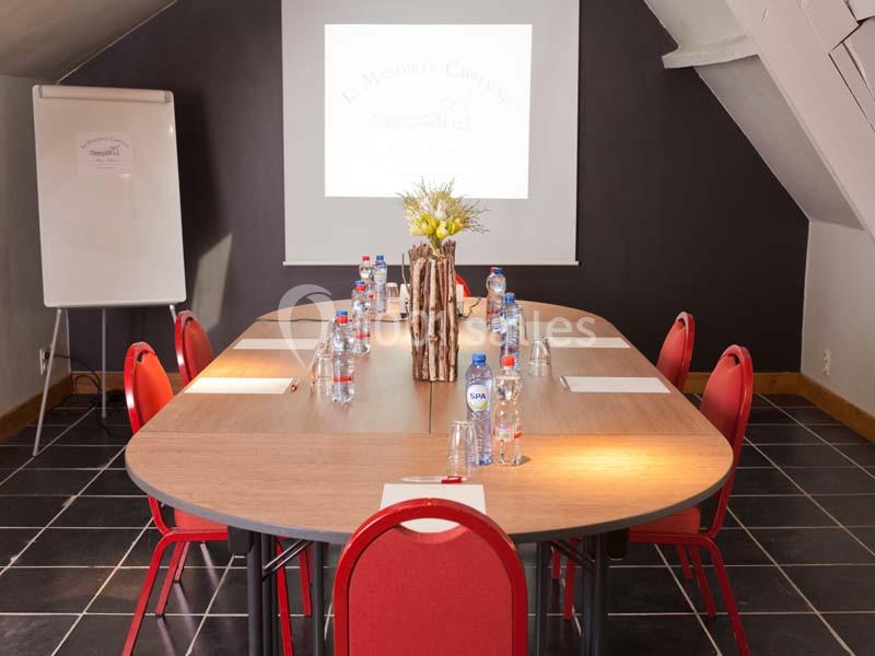 Salle de réunion avec table ovale, chaises rouges, bloc-notes, bouteilles d'eau et écran de projection.