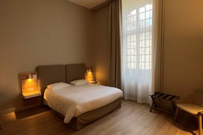 Chambre avec lit double, chevets éclairés, fauteuil, porte-bagages et grande fenêtre avec rideaux translucides.