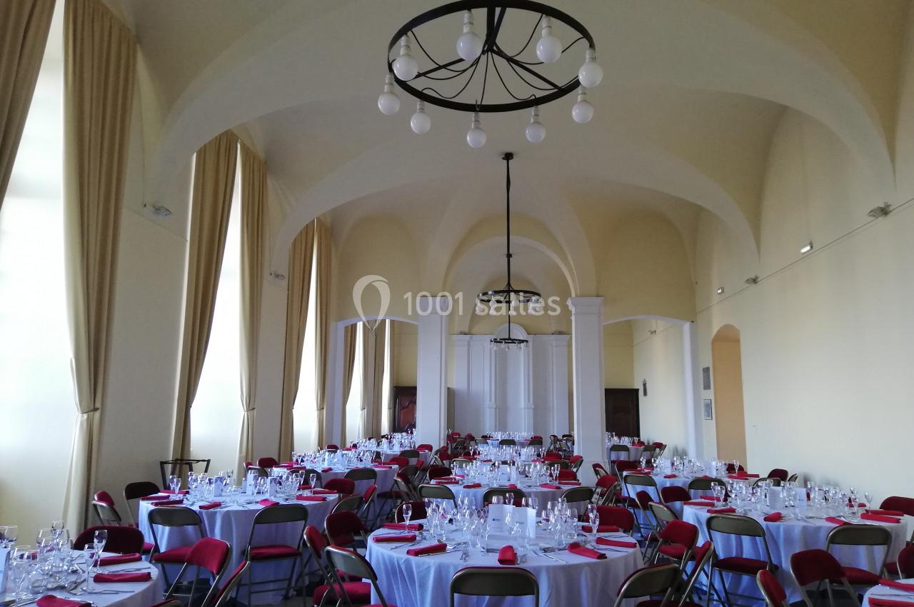 Salle de réception lumineuse avec tables rondes dressées, chaises rouges et lustres suspendus.