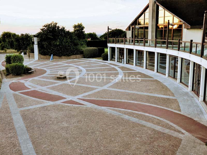 Cour pavée avec motifs géométriques, bordée par un bâtiment vitré et entourée de végétation.