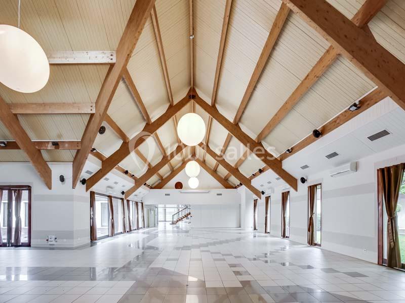 Grande salle lumineuse avec plafond en bois à poutres apparentes, carrelage clair et larges baies vitrées.