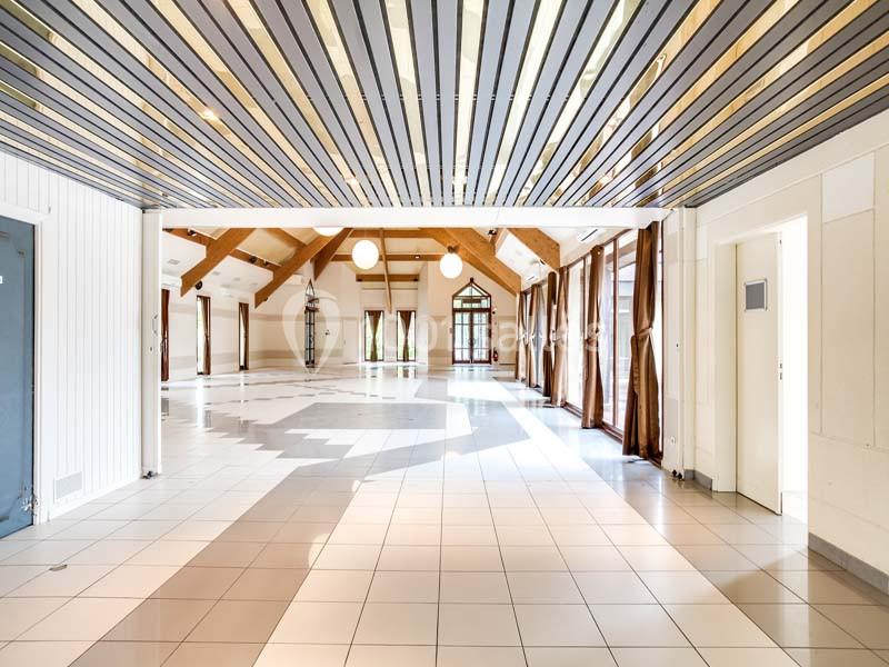 Salle lumineuse avec plafond en lames de bois, grandes baies vitrées, carrelage clair et rideaux marron.