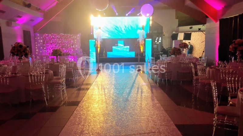 Salle de réception décorée avec des tables rondes, des chaises transparentes et un éclairage coloré autour d'une scène…
