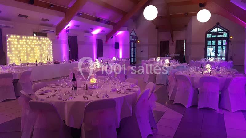 Salle de réception décorée avec des tables rondes dressées, éclairage violet et guirlandes lumineuses au mur.