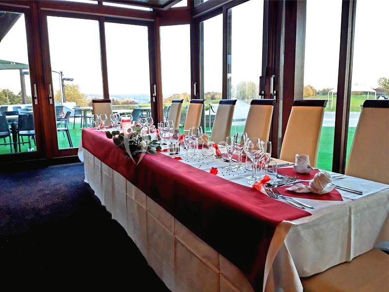 Table dressée avec nappes blanches et rouges, couverts élégants et décorations florales, dans une salle lumineuse avec vue…