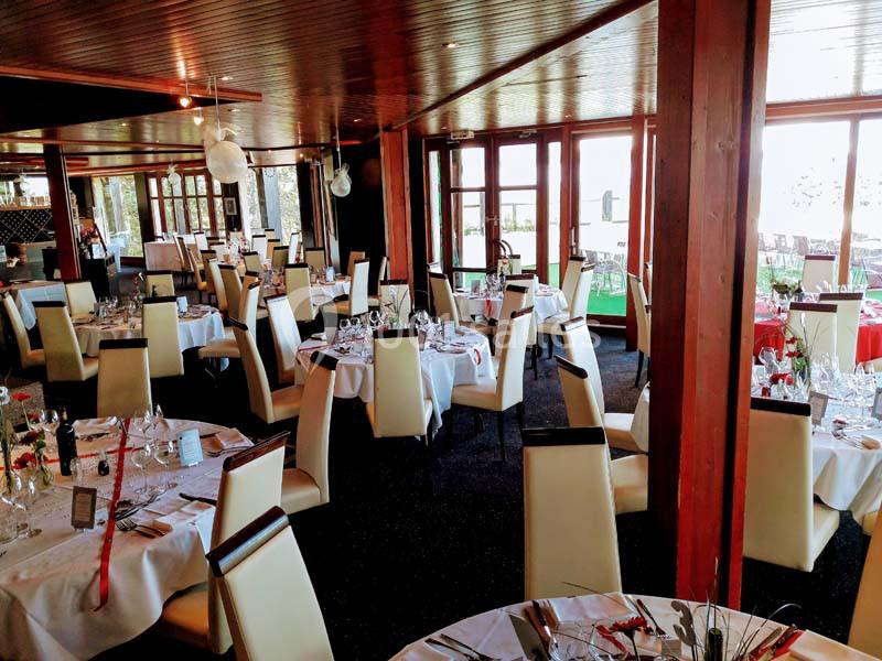 Salle de restaurant lumineuse avec tables dressées, chaises blanches et grandes baies vitrées donnant sur l'extérieur.