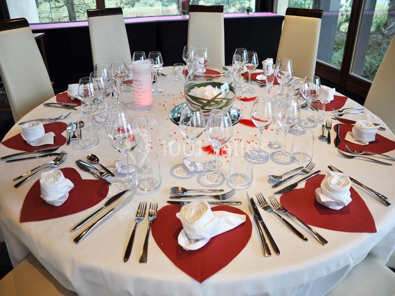 Table ronde élégamment dressée avec nappes blanches, serviettes pliées en roses, décorations rouges et verres alignés.