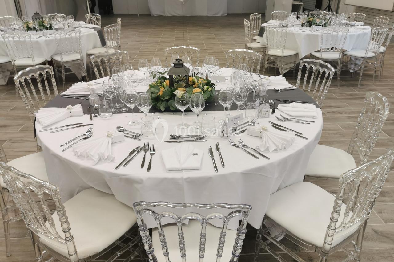 Table ronde élégamment dressée avec nappes blanches, couverts, verres et centre de table floral, entourée de chaises…