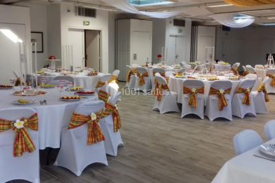 Salle de réception décorée avec des tables rondes, nappes blanches et chaises ornées de nœuds colorés.