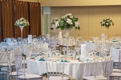 Salle lumineuse avec tables rondes dressées, nappes blanches, chaises rouges et décorations florales élégantes.