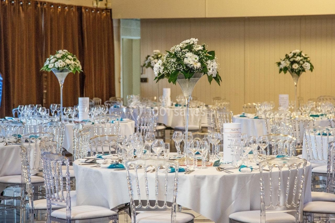 Salle de réception décorée avec des tables rondes, nappes blanches, chaises transparentes et centres de table floraux.