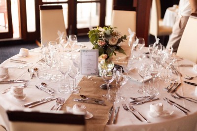 Salle lumineuse avec tables rondes dressées, nappes blanches, chaises rouges et décorations florales élégantes.