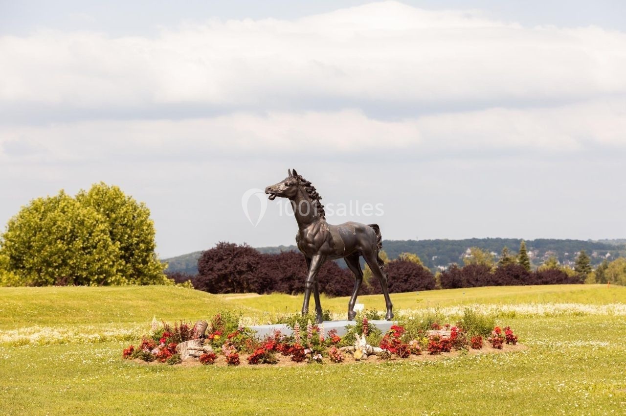 Statue d'un poulain en bronze entourée de fleurs sur une pelouse verdoyante avec des arbres et un ciel nuageux en arrière…