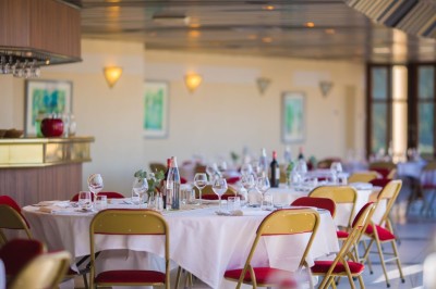 Salle lumineuse avec tables rondes dressées, nappes blanches, chaises rouges et décorations florales élégantes.