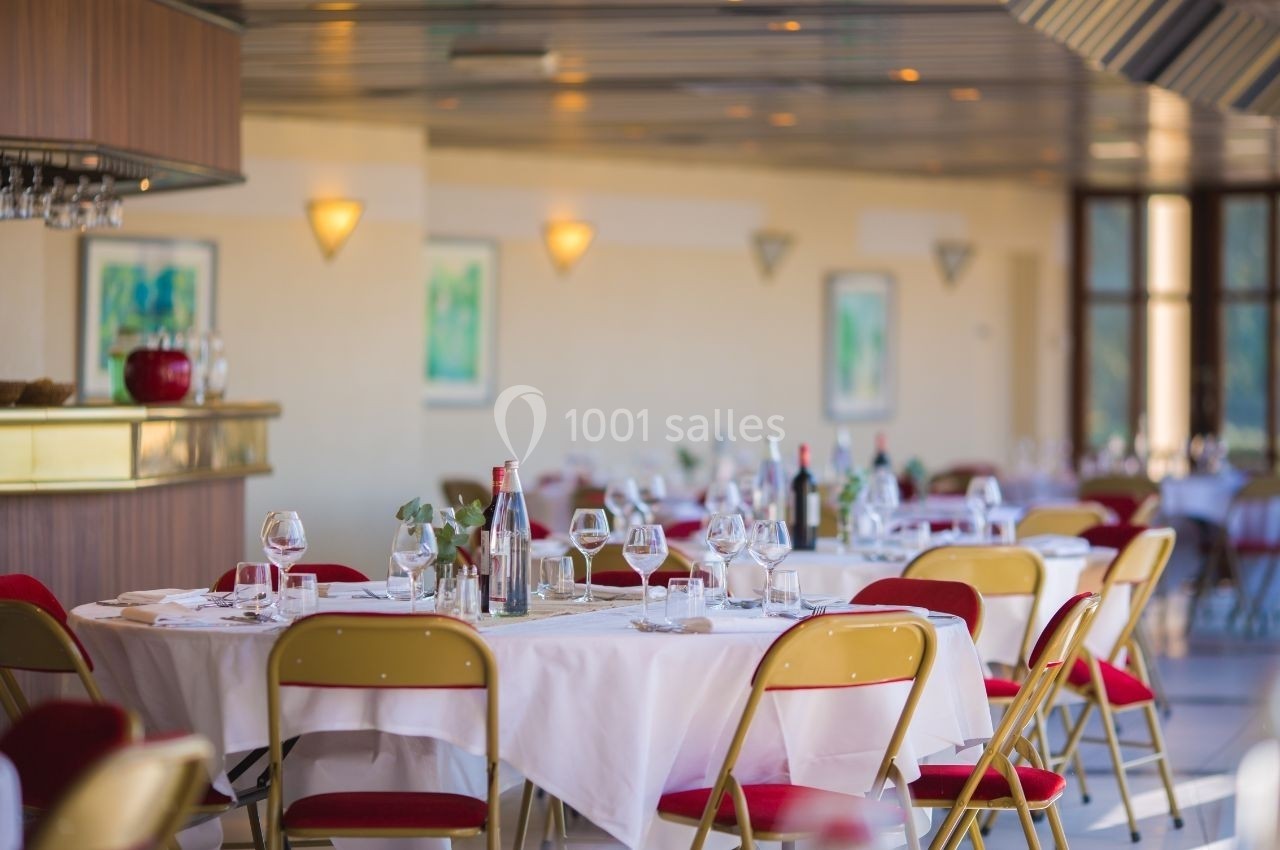 Salle de restaurant lumineuse avec tables dressées, nappes blanches, chaises rouges et bar en arrière-plan.