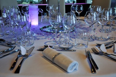 Salle lumineuse avec tables rondes dressées, nappes blanches, chaises rouges et décorations florales élégantes.