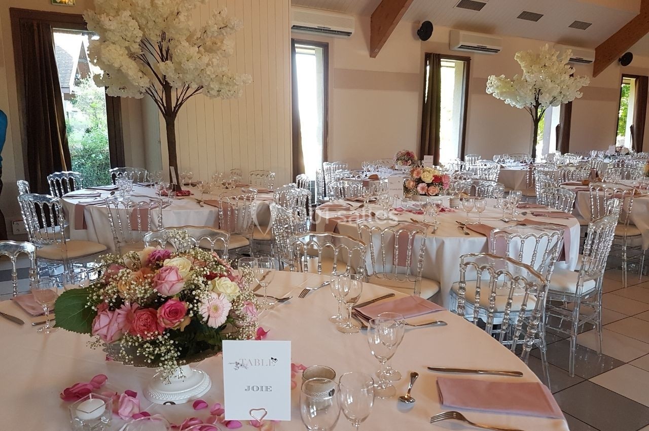 Salle de réception décorée pour un mariage avec tables rondes, nappes blanches, chaises transparentes et arrangements…