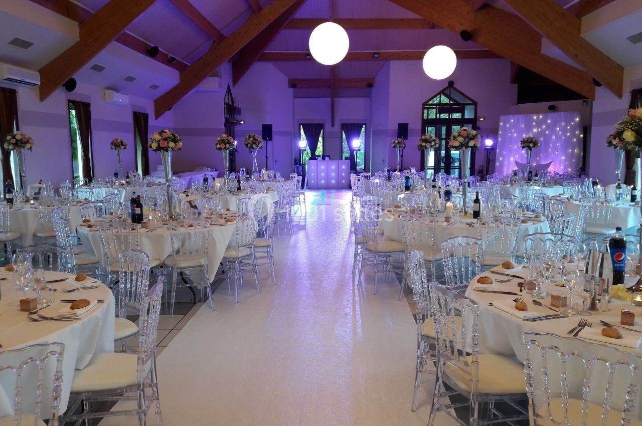 Salle de réception décorée avec des tables rondes dressées, chaises transparentes et éclairage tamisé.