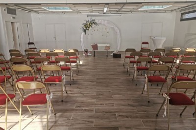 Salle lumineuse avec tables rondes dressées, nappes blanches, chaises rouges et décorations florales élégantes.