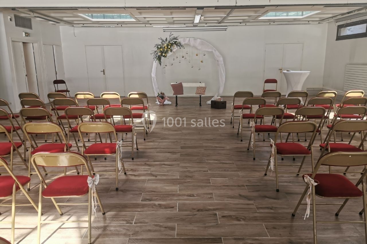Salle lumineuse aménagée pour une cérémonie, avec des chaises rouges alignées et une arche décorée de fleurs.
