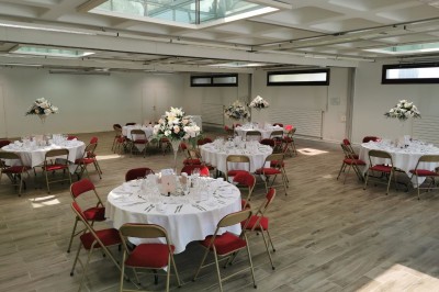 Salle lumineuse avec tables rondes dressées, nappes blanches, chaises rouges et décorations florales élégantes.