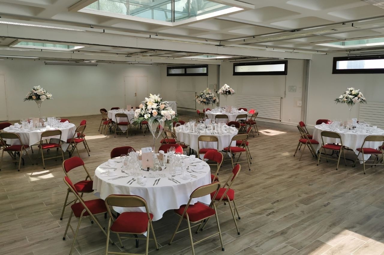 Salle lumineuse avec tables rondes dressées, nappes blanches, chaises rouges et décorations florales élégantes.