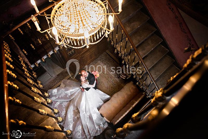 Vue plongeante sur un couple de mariés enlacés au pied d'un escalier en bois, sous un lustre élégant.