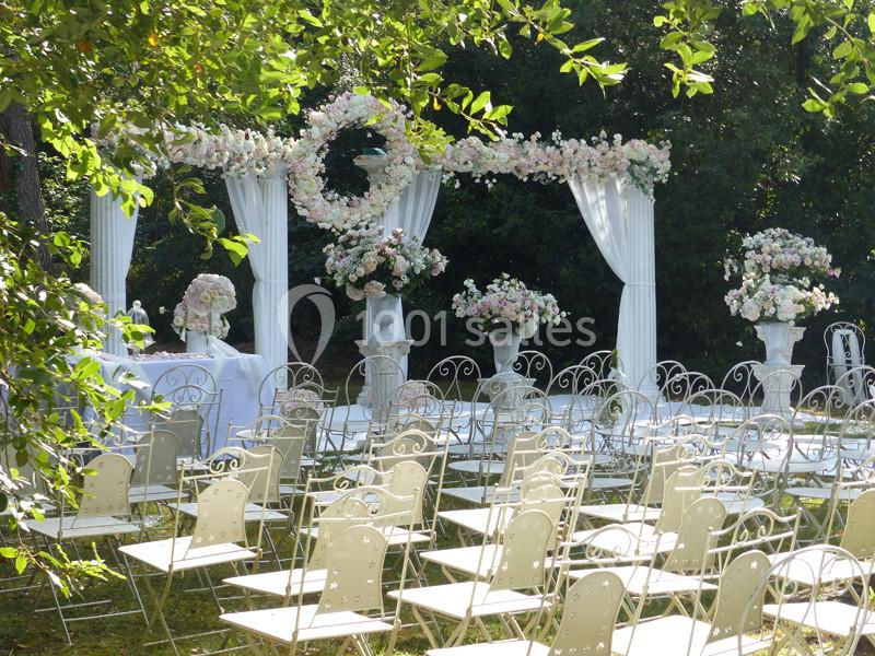 Chaises blanches alignées devant une arche décorée de fleurs blanches et roses dans un jardin ensoleillé.