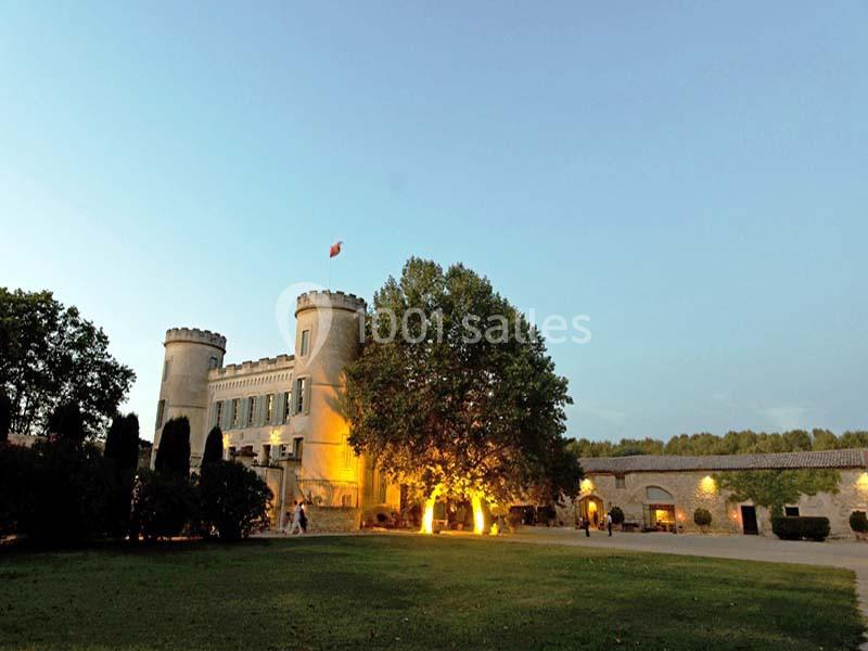 Château éclairé au crépuscule, entouré d'arbres et d'une pelouse, avec un drapeau flottant au sommet.