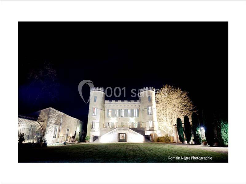 Façade éclairée d'un château avec deux tours rondes, entouré d'arbres et d'une pelouse, vue de nuit.