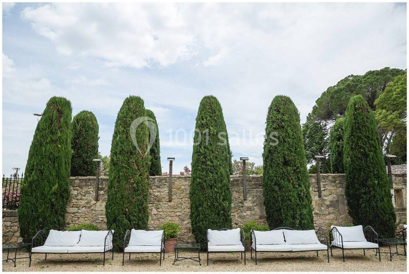 Six cyprès alignés devant un mur en pierre, avec des bancs en fer forgé et coussins blancs disposés en dessous.