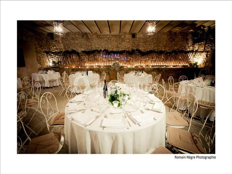 Salle de réception avec tables rondes dressées, chaises en fer forgé et murs en pierre éclairés par des lumières tamisées.