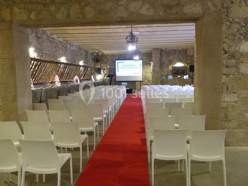 Salle en pierre avec des rangées de chaises blanches, un tapis rouge central et un écran de projection en fond.