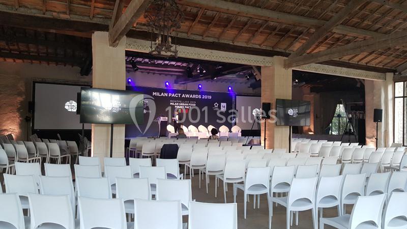Salle de conférence avec des rangées de chaises blanches, une scène éclairée et des écrans affichant des informations.