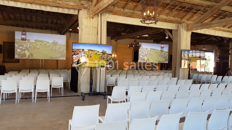 Salle de conférence avec des rangées de chaises blanches et plusieurs écrans affichant une scène en extérieur.