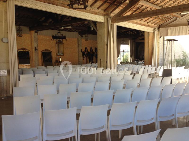 Salle de réception avec rangées de chaises blanches alignées, murs en pierre et poutres apparentes au plafond.